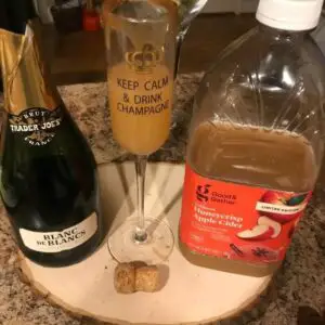 A glass of easy cozy apple cider mimosas drink between a bottle of champagne and gallons of apple cider.