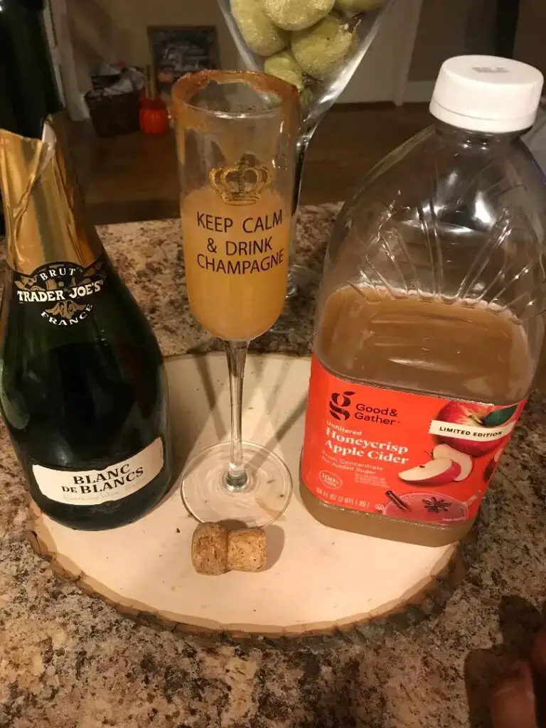 cozy apple cider mimosas drink between a bottle of champagne and apple cider gallons.