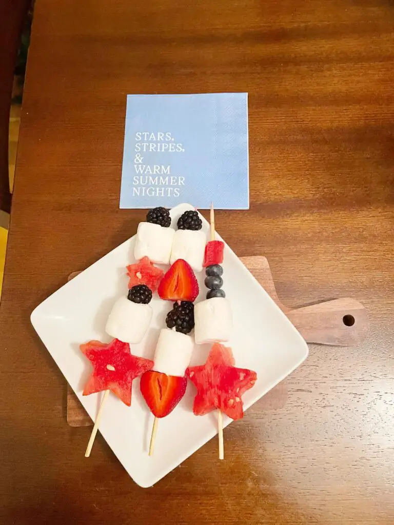 Star shaped watermelon, marshmallows, strawberries, blueberries, and blackberries are arranged alternatingly on a kabob stick and placed on a square plate.