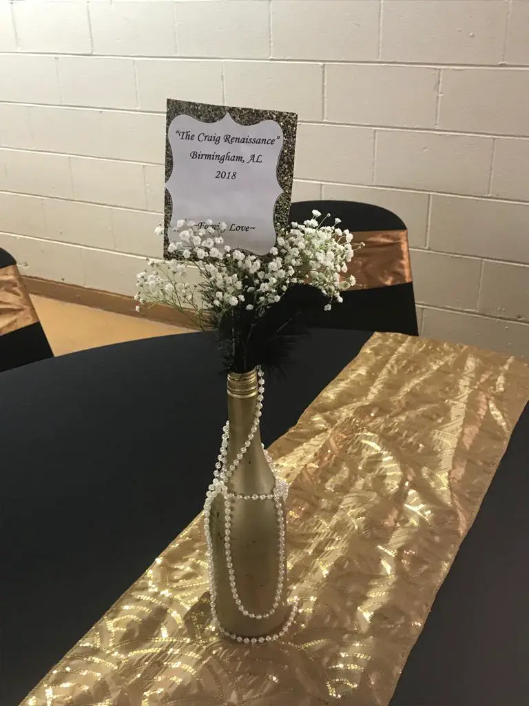 Round table decorated with a bottle with white flowers and a note. Black chairs with golden bands in the middle.