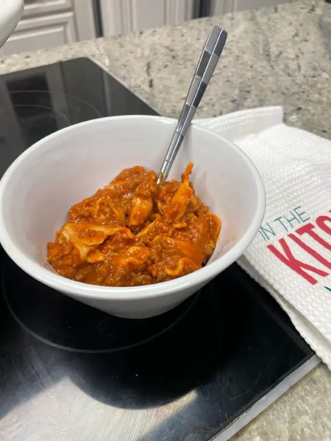 comforting and hearty lasagna soup in a white bowl with a silver spoon placed on a black stovetop next to a branded towel from KMarie in the Kitchen