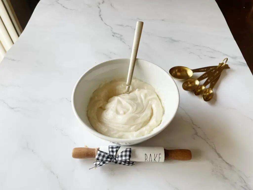 easy cream cheese icing in a white bowl with decorative wooden dough roller on a white marble countertop