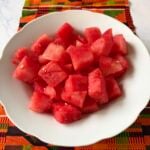 A white bowl filled with freshly cubed, juicy red watermelon pieces, set against a vibrant orange, green, and black geometric patterned Kente cloth placemat.