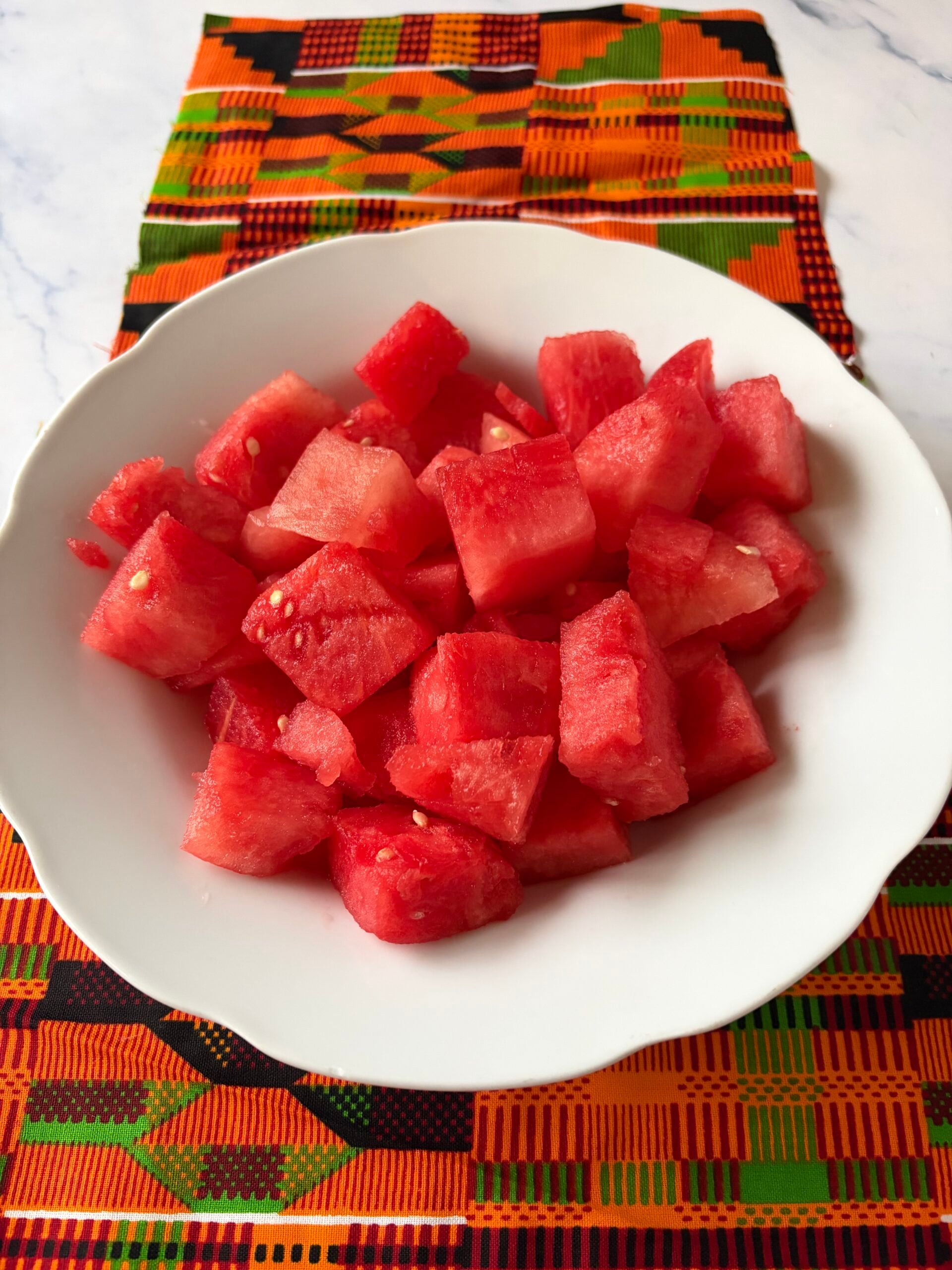 A white bowl filled with freshly cubed, juicy red watermelon pieces, set against a vibrant orange, green, and black geometric patterned Kente cloth placemat.