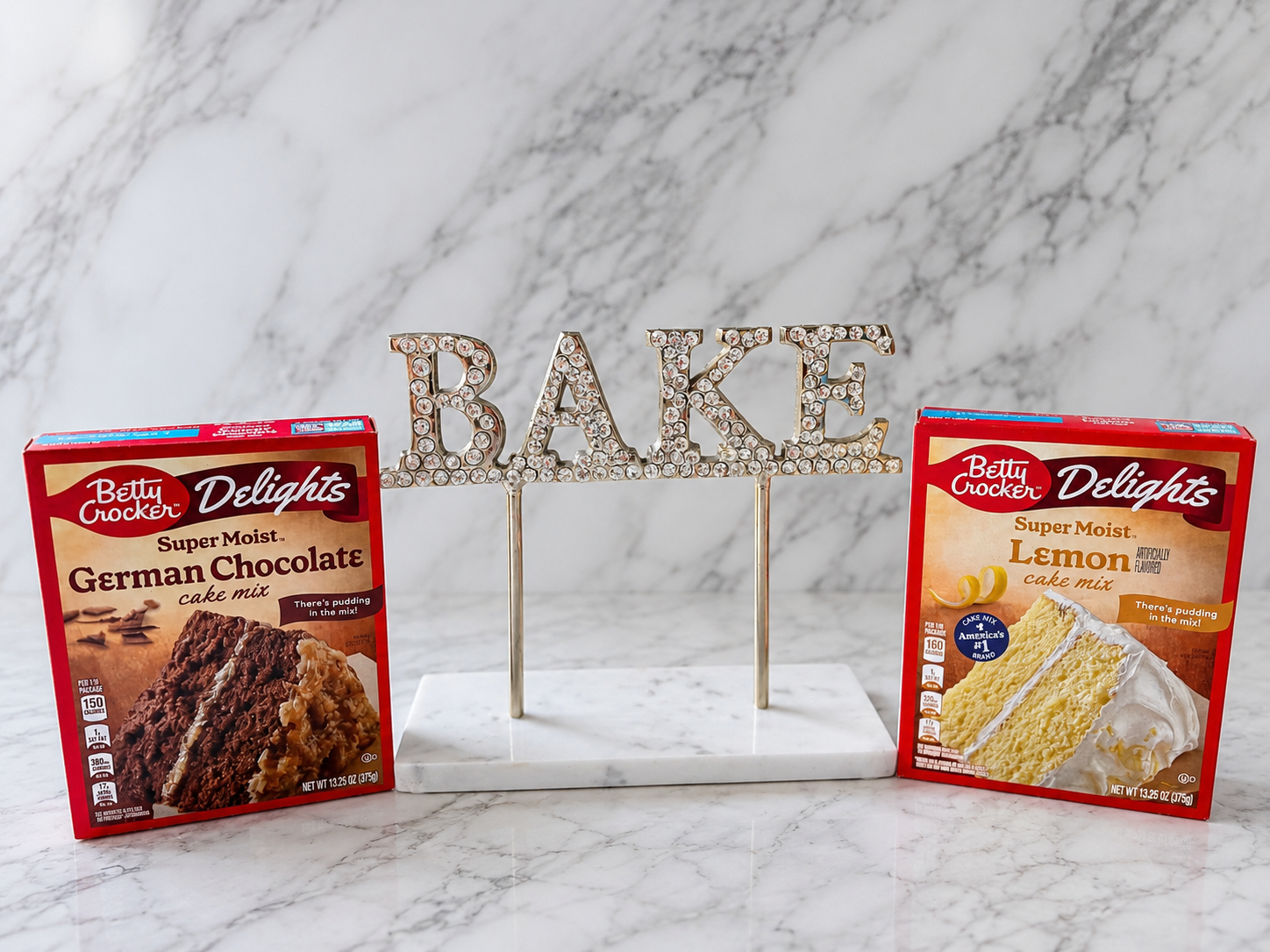 Betty Crocker German chocolate and lemon box cake mixes displayed beside a sparkling BAKE sign on a white marble background.
