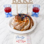 Patriotic Funfetti Bundt cake with vanilla glaze served with red drinks and Fourth of July decorations on a white marble background.